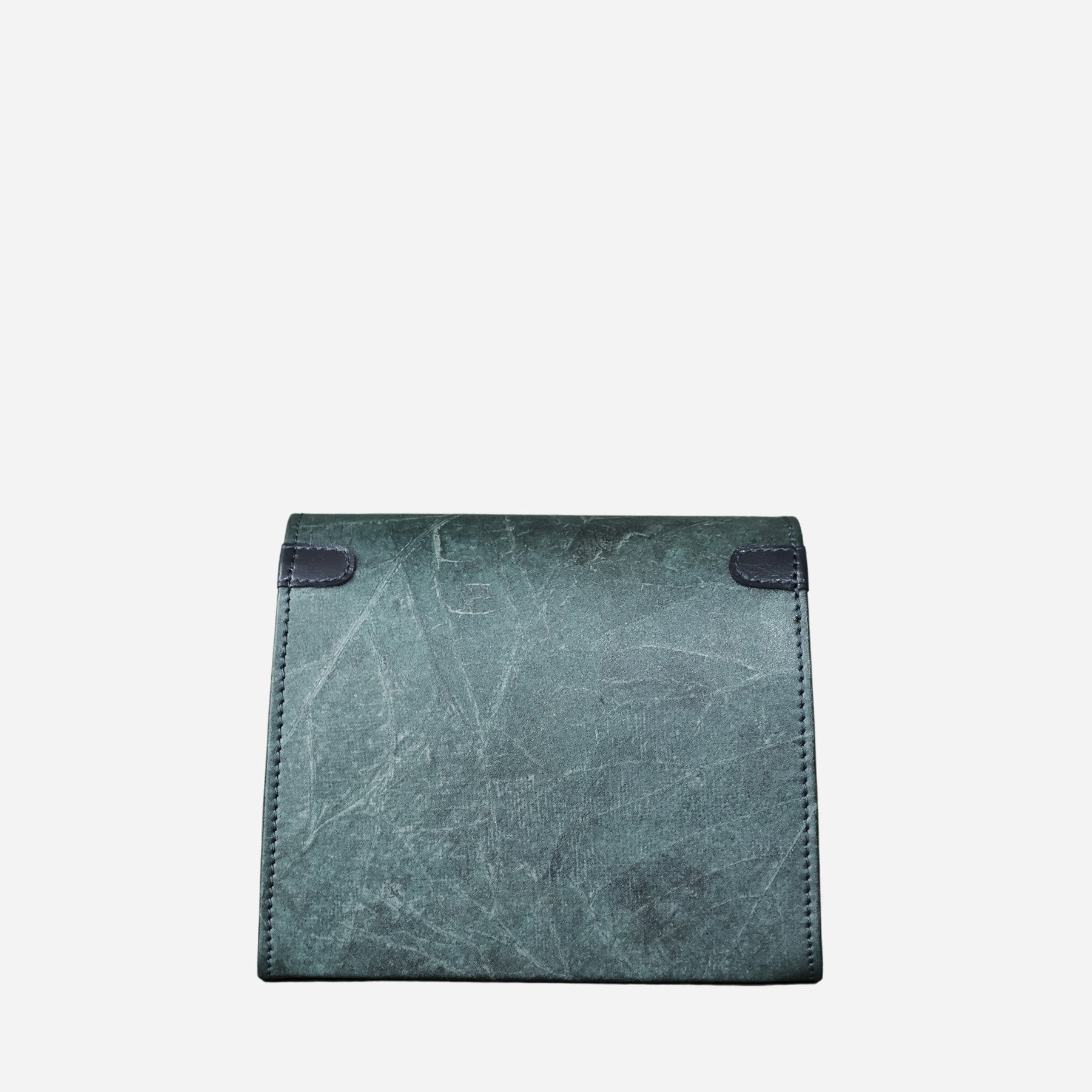 A back viewed of the blue dianne vegan crossbody bag by Thamon with a leaf textured surface, closed flap, and dark blue edging, displayed against a white background. The bag has a boxy shape and a minimalist design