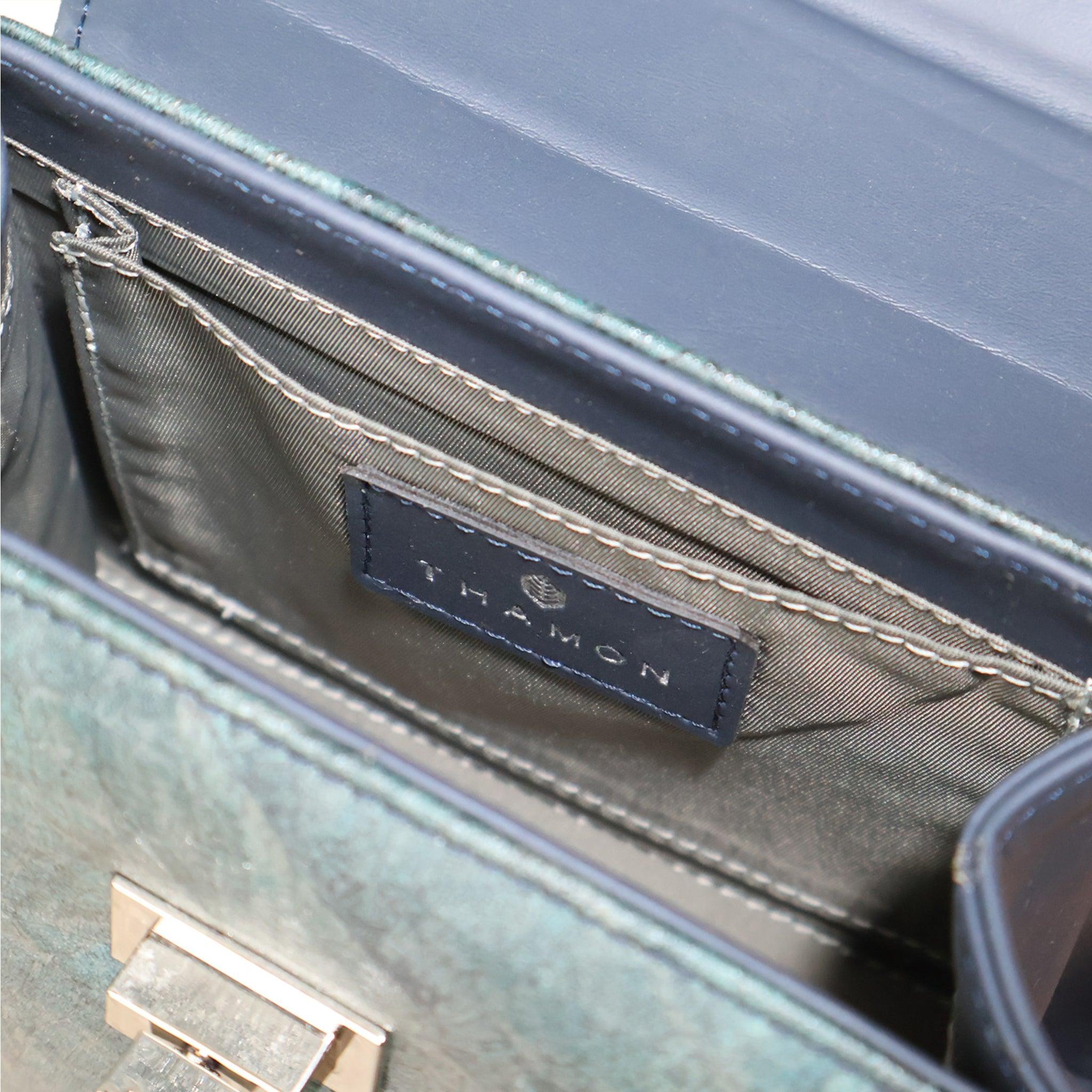 Close-up view of the interior of a metallic green handbag, showing the open main compartment. The image highlights a small internal slip pocket with a dark leather patch featuring the brand name "THAMON." The bag's shiny silver-tone twist-lock closure is also visible on the front flap. The interior is lined with a dark gray fabric.