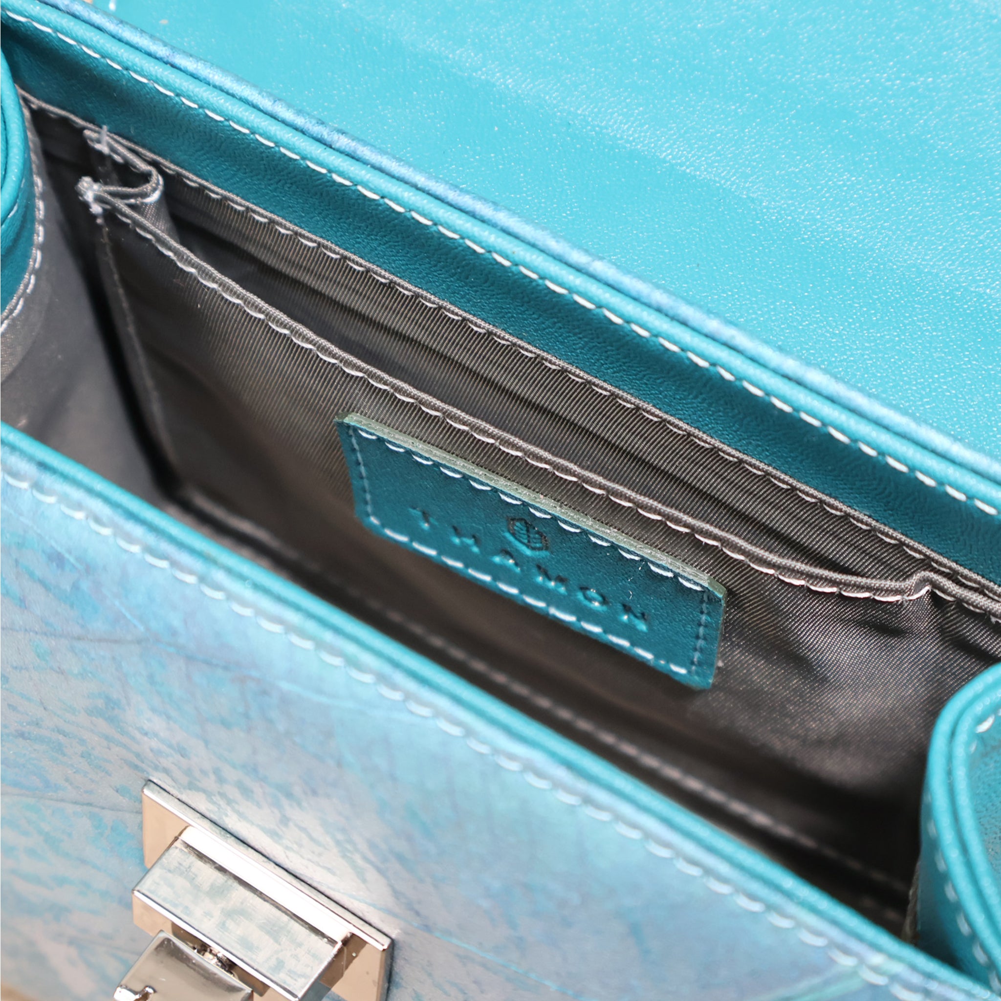 Close-up view of the interior of a teal-colored handbag, showing the open main compartment. The image highlights a small internal slip pocket with a matching leather patch. The bag's shiny silver-tone twist-lock closure is also visible on the front flap. The interior is lined with a dark gray fabric and features exposed white stitching.