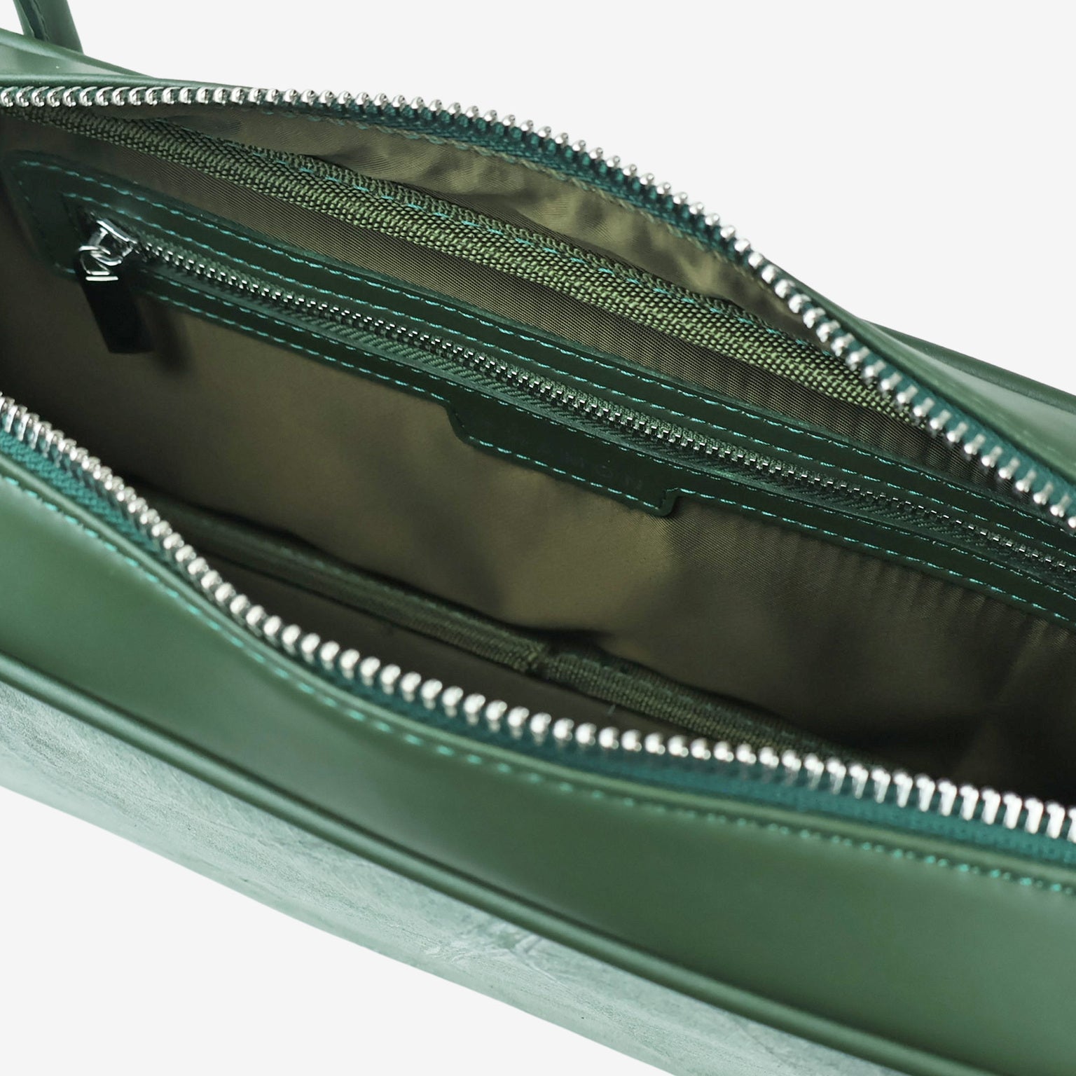 A close-up, high-angle view of the open zipper of a dark green vegan leather bag, revealing its olive-green fabric lining and an internal zippered pocket.