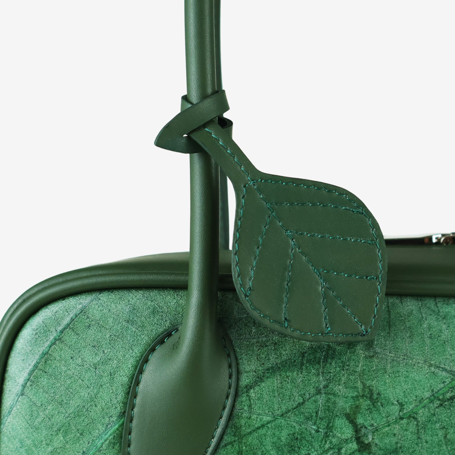 A close-up of a dark green vegan leather shoulder bag, showcasing a detailed leaf-shaped charm with stitched veins hanging from the handle.