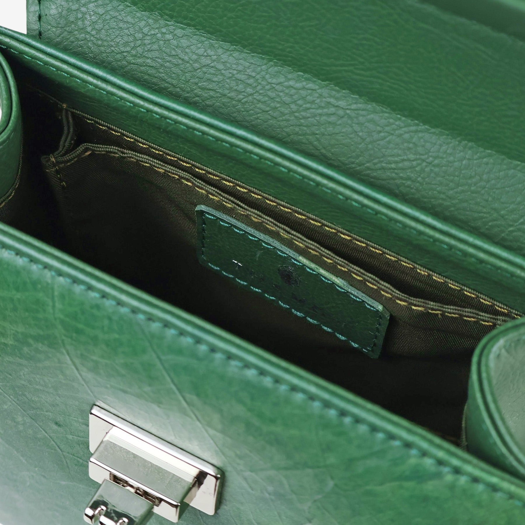 Close-up view of the interior of a deep green handbag, showing the open main compartment. The image highlights a small internal slip pocket with a matching leather patch. The bag's shiny silver-tone twist-lock closure is also visible on the front flap. The interior is lined with a dark green fabric and features exposed stitching.