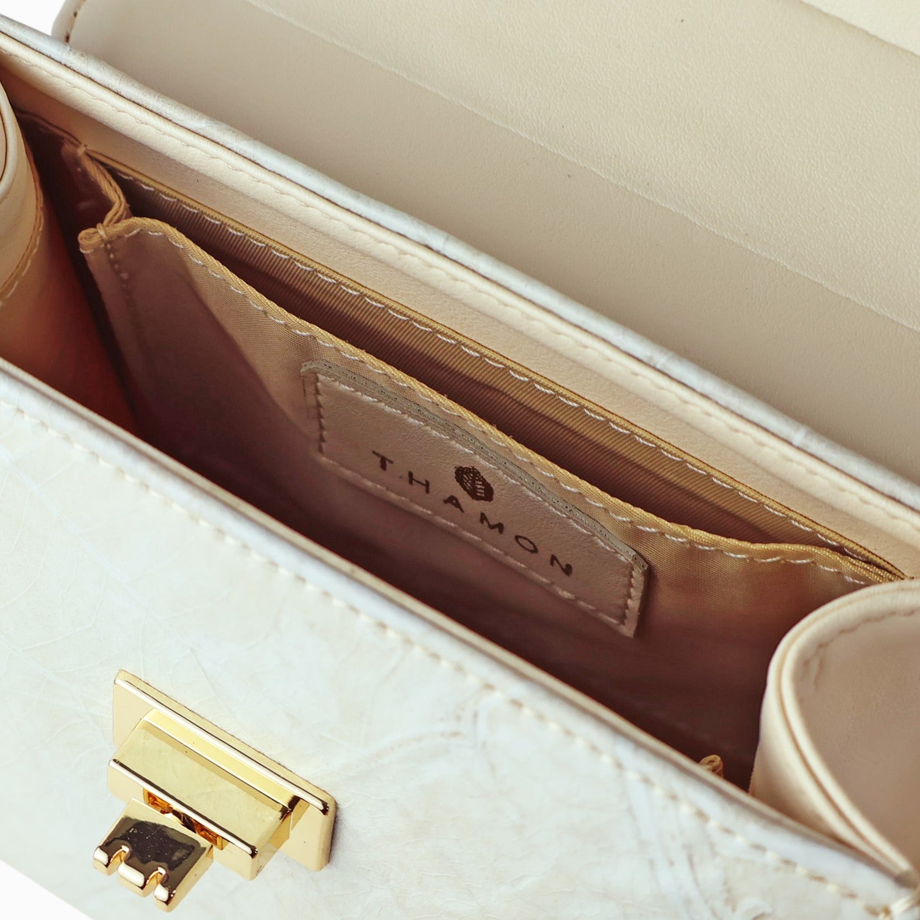 Close-up view of the interior of a cream-colored handbag, showing the open main compartment. The image highlights a small internal slip pocket with a leather patch featuring the brand name "T.HAMON." The bag's gold-tone twist-lock closure is also visible on the front flap. The interior is lined with a beige fabric.