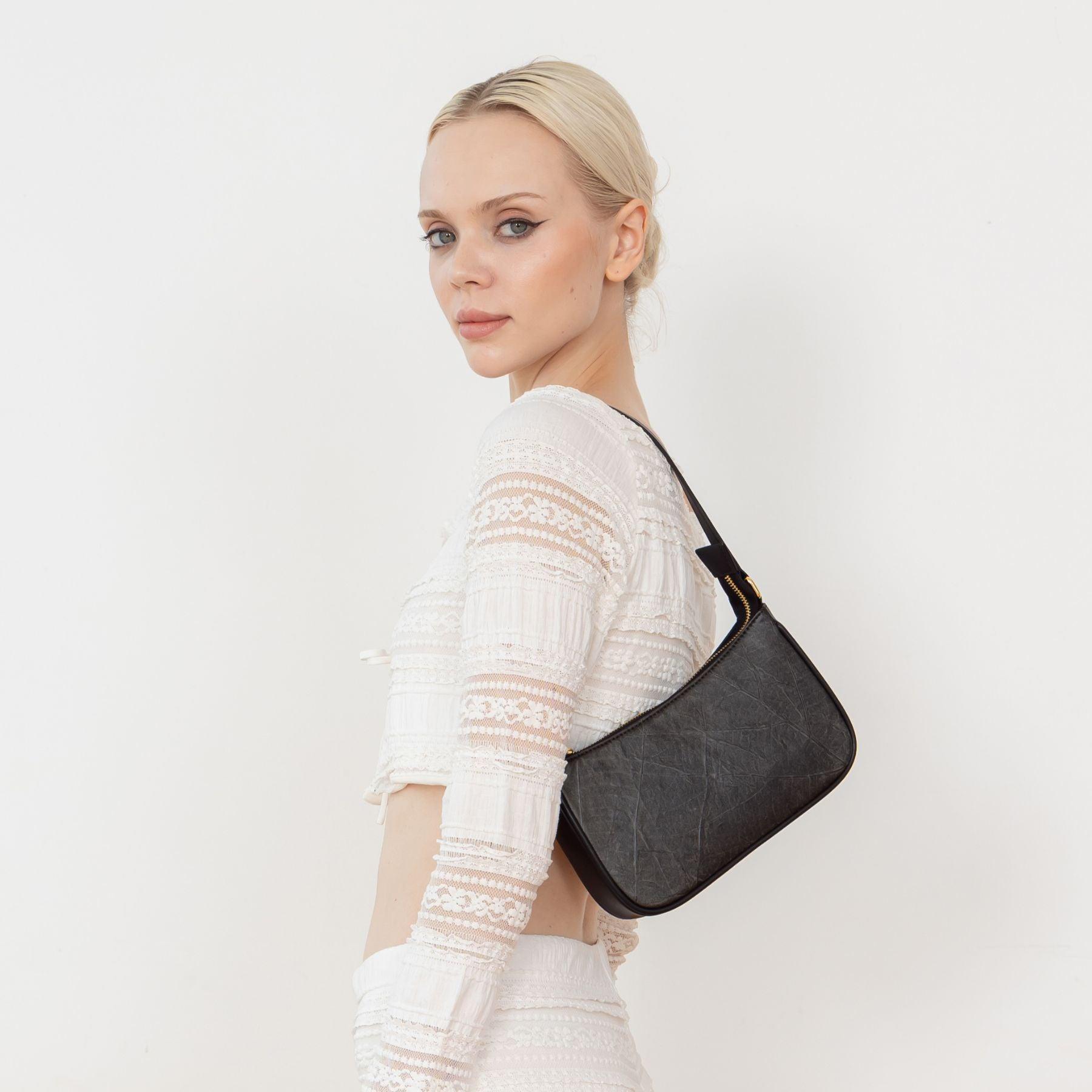 A woman wearing a white blouse and white pants, showcasing the Mila Black Vegan Shoulder Bag with a detailed leaf pattern