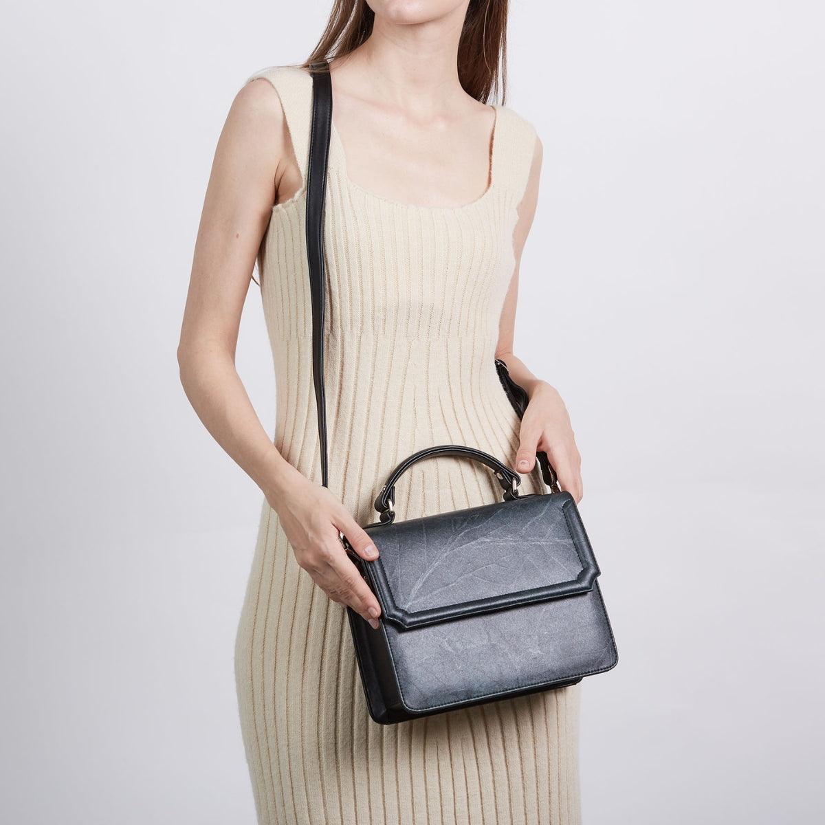 Model wearing cream dress holding black Camden crossbody bag with top handle and adjustable strap, made from Leaftex