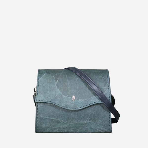 A blue Dianne box-style vegan crossbody bag with a leaf imprint texture on the front, a dark blue strap, and a small leaf logo metallic emblem in the center, against a plain white background.