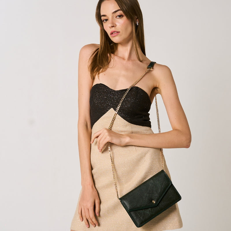 A model with a sleek hairstyle wears a strapless black and gold shimmer mini dress, accessorized with a forest green vegan leather pouch bag with a gold chain, against a simple white background.