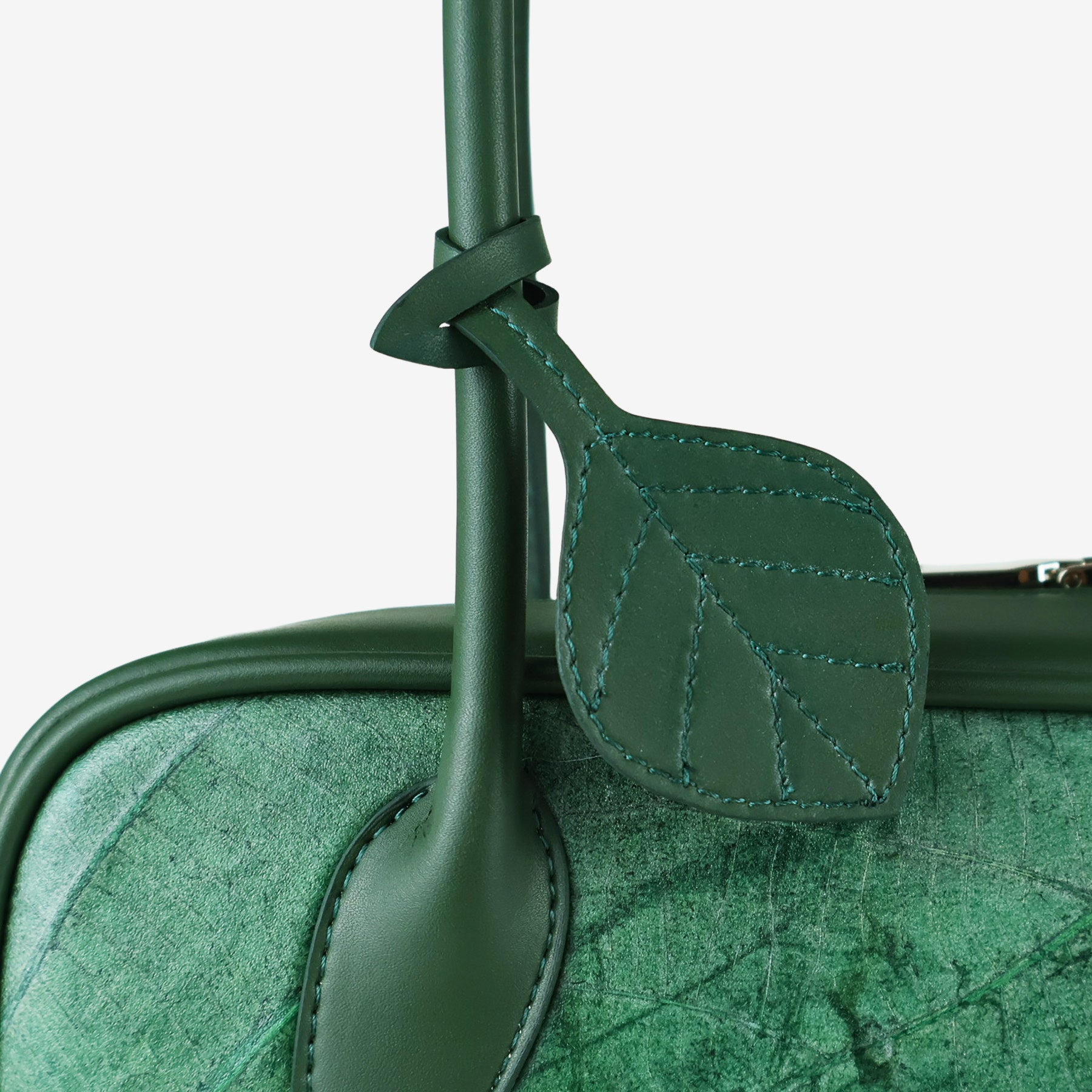 A close-up of a dark green vegan leather shoulder bag, showcasing a detailed leaf-shaped charm with stitched veins hanging from the handle.