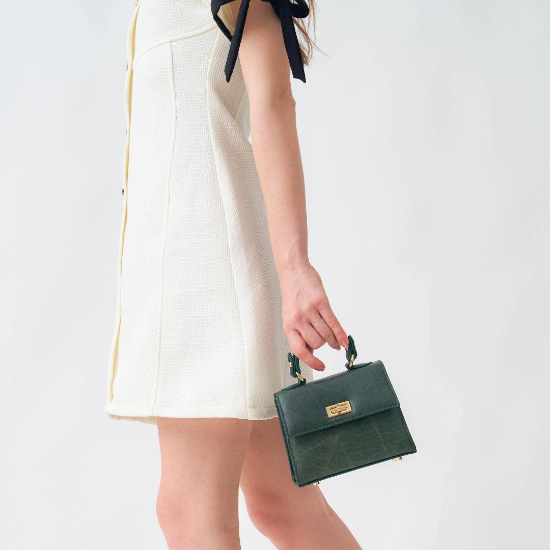 Model wearing a cream dress, holding the Thamon Kylie Mini Bag in Forest Green Leaf Leather, highlighting the bag's chic and compact design.
