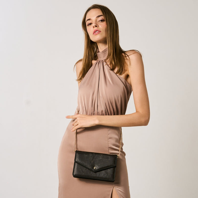 A poised young woman in a pale pink halter neck dress with a high slit, holding a black vegan leather pouch bag with a gold chain strap, set against a neutral backdrop