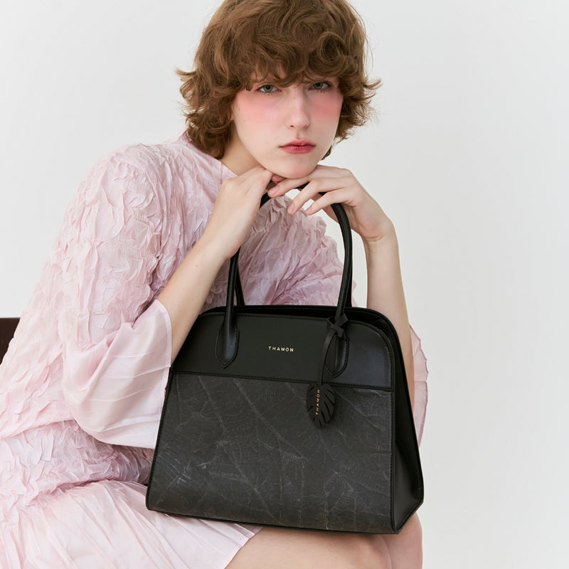 Woman seated holding the Sloane Tote Bag in black, showcasing the elegant leaf pattern and sophisticated design.