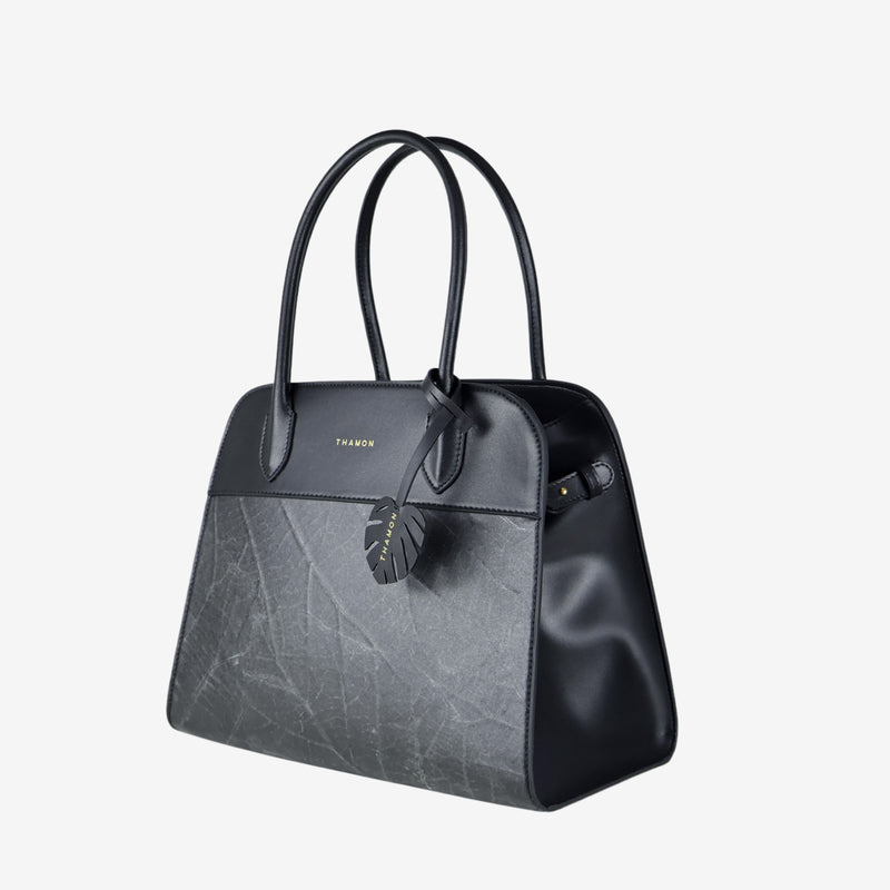 Side view of the Sloane tote bag in black vegan leather with a pressed leaf pattern, featuring sturdy top handles, a structured silhouette, and a decorative leaf-shaped tag attached to the front handle.