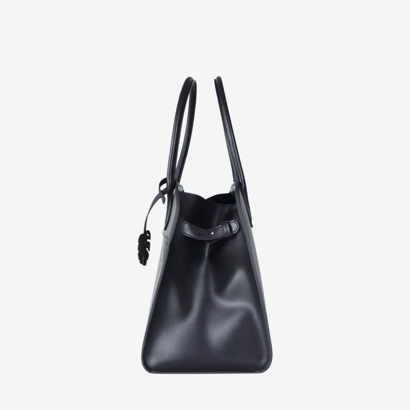 Side profile of the Sloane tote bag in black vegan leather, featuring a smooth, structured surface, sturdy top handles, and a minimal design with a decorative leaf-shaped tag visible near the front.