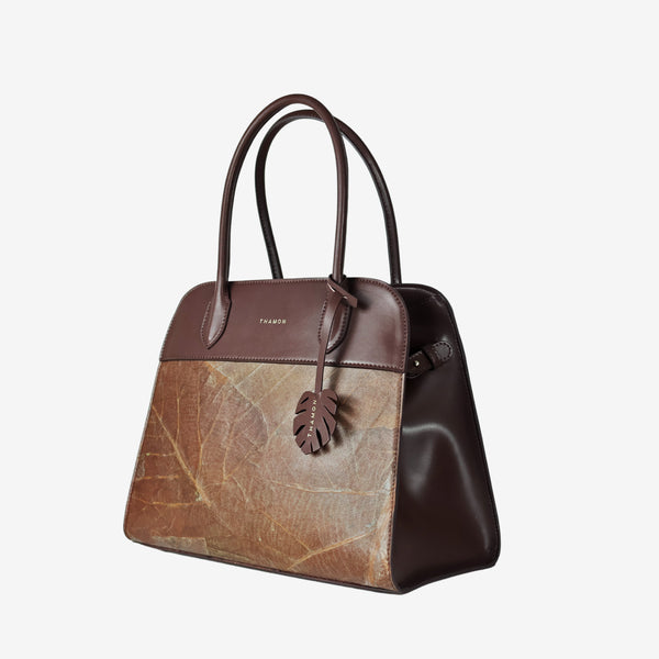 Side view of the Sloane tote bag in spice brown vegan leather with a pressed leaf pattern, featuring sturdy top handles, a structured silhouette, and a decorative leaf-shaped tag attached to the front handle.