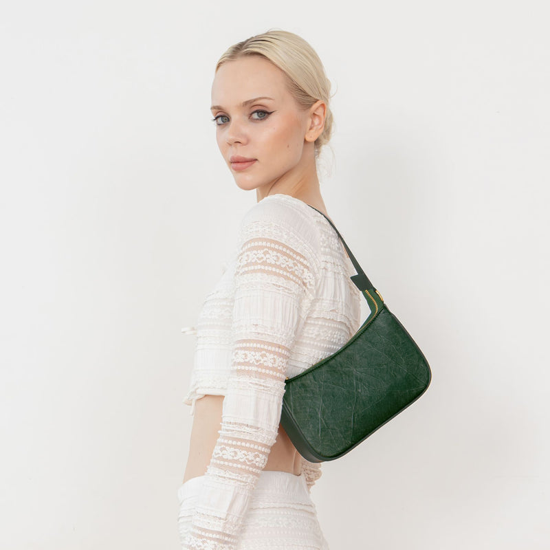 A woman wearing a white blouse and white pants, showcasing the Mila forest green Vegan Shoulder Bag with a detailed leaf pattern