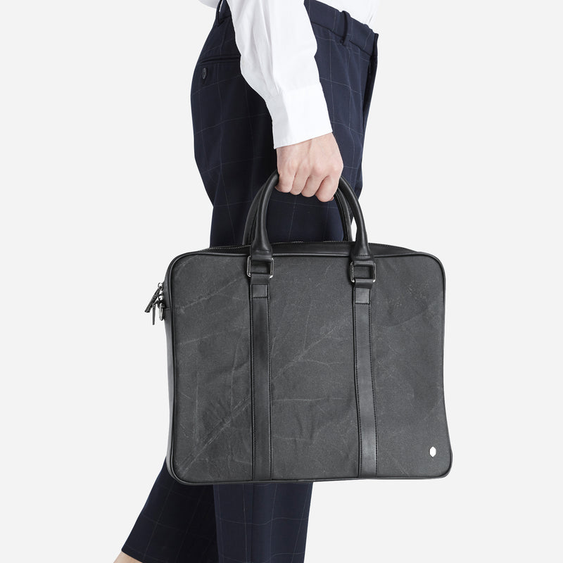A person holding a black leaf-patterned leather briefcase by Thamon with top handles against a navy checkered trouser and white shirt background.