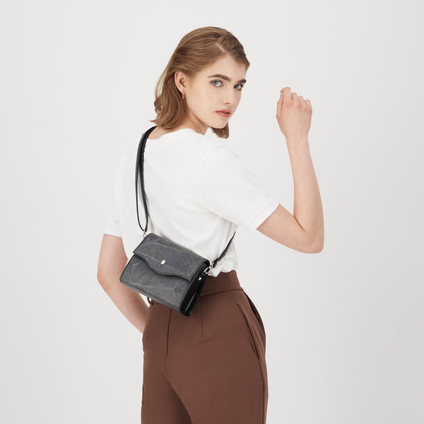 Model wearing a Thamon London black vegan leather box bag, positioned to display the bag's structured design and leaf leather texture against a white top and brown trousers, with the bag's strap over her shoulder.