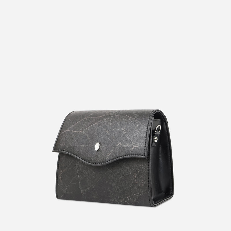 Side view of Thamon London's black leaf leather box bag showing the distinctive textured surface, the curved flap closure with a silver-toned snap button, and the sleek side profile with visible stitching details.