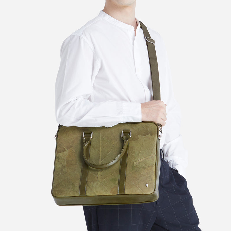 A side profile of a man wearing a white shirt and navy blue checkered pants, holding a Cambridge-style briefcase made from olive leaf leather. The briefcase has an olive green leaf pattern, rounded handles, and an adjustable olive green shoulder strap. The product is positioned to highlight the eco-friendly material and stylish design, targeted for a professional setting.