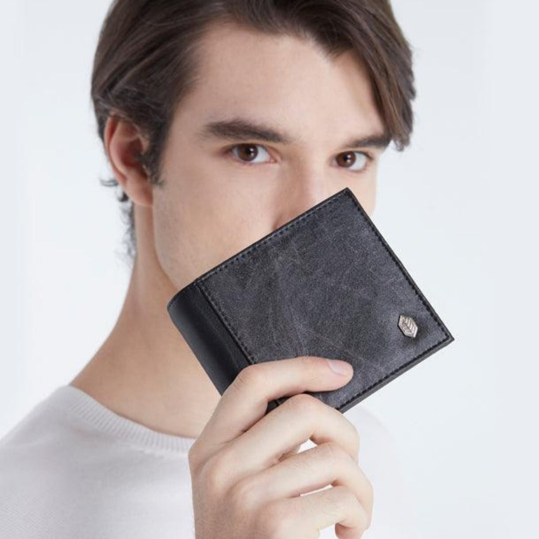 Model wearing a white crewneck sweater holding a black oliver vegan leather wallet near his face, showcasing its smooth texture and silver leaf logo. The minimalist background emphasizes the wallet's design and craftsmanship.