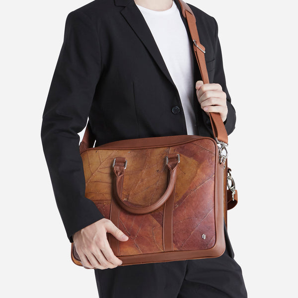 A man in a black suit holding a Thamon spice brown leaf leather Cambridge briefcase with a shoulder strap.