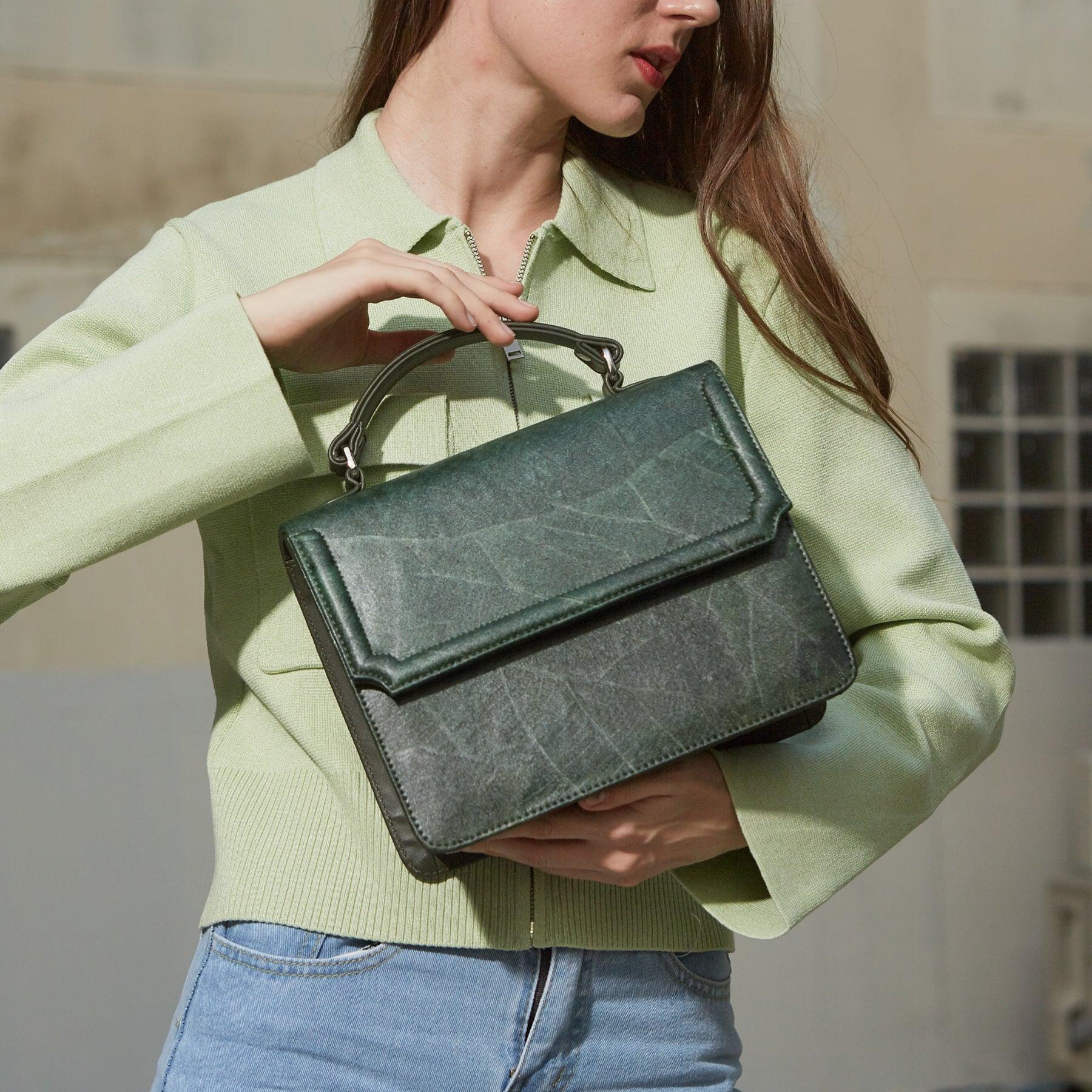 Model in light green jacket holding Camden crossbody bag in forest green Leaftex with top handle and flap design