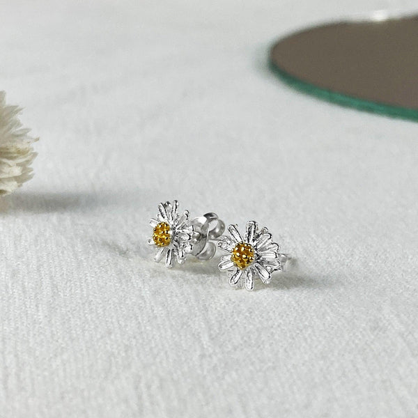 Silver daisy flower stud earrings with a gold center, handcrafted by Thamon, displayed on a soft white fabric background.